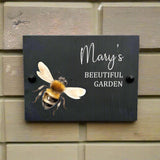 Personalised Slate Garden House Sign Featuring a Printed Honey Bee Illustration. Custom Rectangular Design With the Name "Mary's" and a message "Beeutiful Garden". Displayed against a Brick Wall Background.